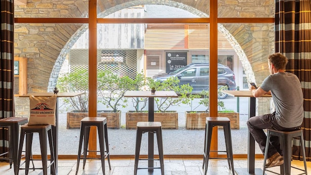 A man sitting at a table inside a cafe with a stone wall and wooden ceiling, overlooking a street with parked cars and a building with a sign. - Apartment in Saint-Lary-Soulan with Pool & Sauna - 6 ppl (Saint-Lary-Soulan)