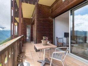 A balcony with a wooden railing, a table with chairs, a basket with plants, and a sliding glass door leading to a room with a TV. - Ski-in/Ski-out apartment with heated pool and parking (Saint-Lary-Soulan)