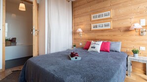 A bedroom with a wooden wall, a bed with grey bedding, red and white pillows, and a small table with a lamp.