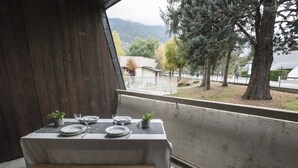 Une table à manger dressée avec des assiettes, des verres et des couverts, placée sur un balcon donnant sur une rue bordée d’arbres et de bâtiments.