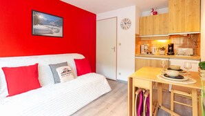 A cozy room with a white sofa, red cushions, a wooden dining table, and a kitchen area with wooden cabinets and a clock.
