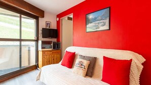 A bedroom with a large window, a bed with white and red bedding, a wooden cabinet with a television and a microwave, and a framed picture on the wall.