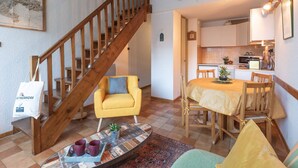 Een gezellig interieur met een houten trap, een gele fauteuil, een eettafel met stoelen en een keukenhoek.