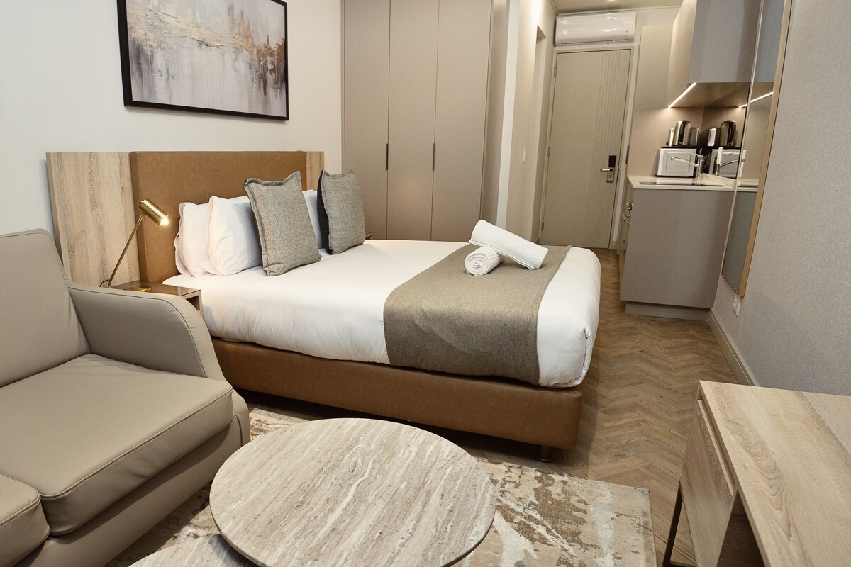Una habitación de hotel moderna con cama, sofá, una mesita de centro redonda y una kitchenette.