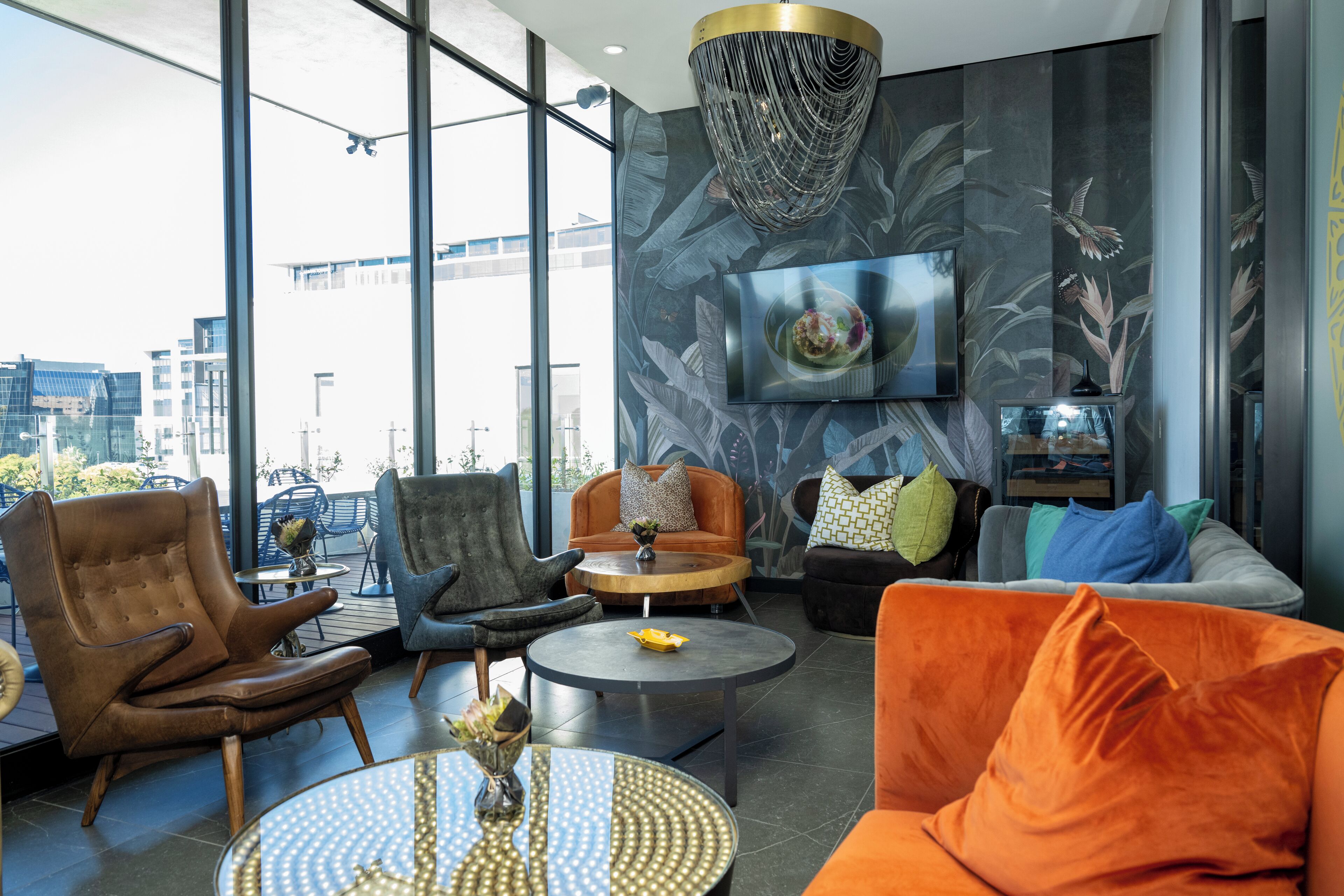 A modern lounge area with leather armchairs, a round coffee table, and a sofa with orange cushions. A television is mounted on the wall, and a decorative chandelier hangs above.