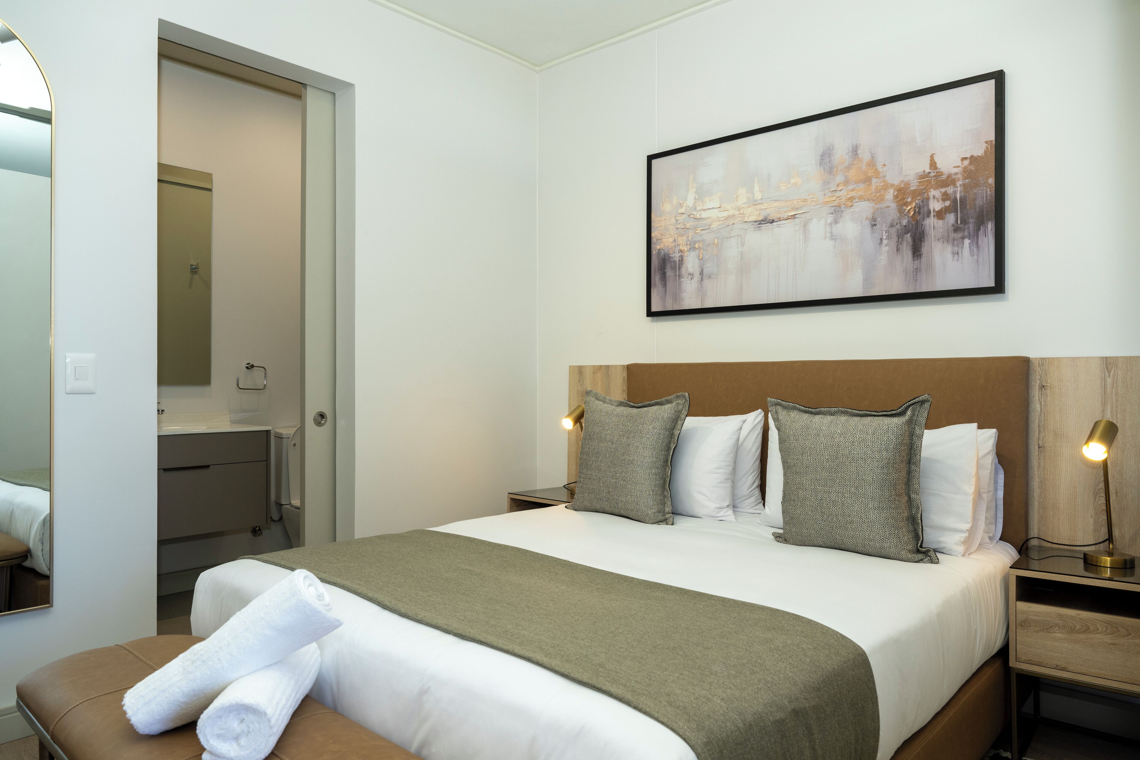 Ein Hotelzimmer mit einem großen Bett, einem Nachttisch mit Lampe, einem Bild an der Wand und einem durchgegangene Tür erkennbaren Badezimmer.