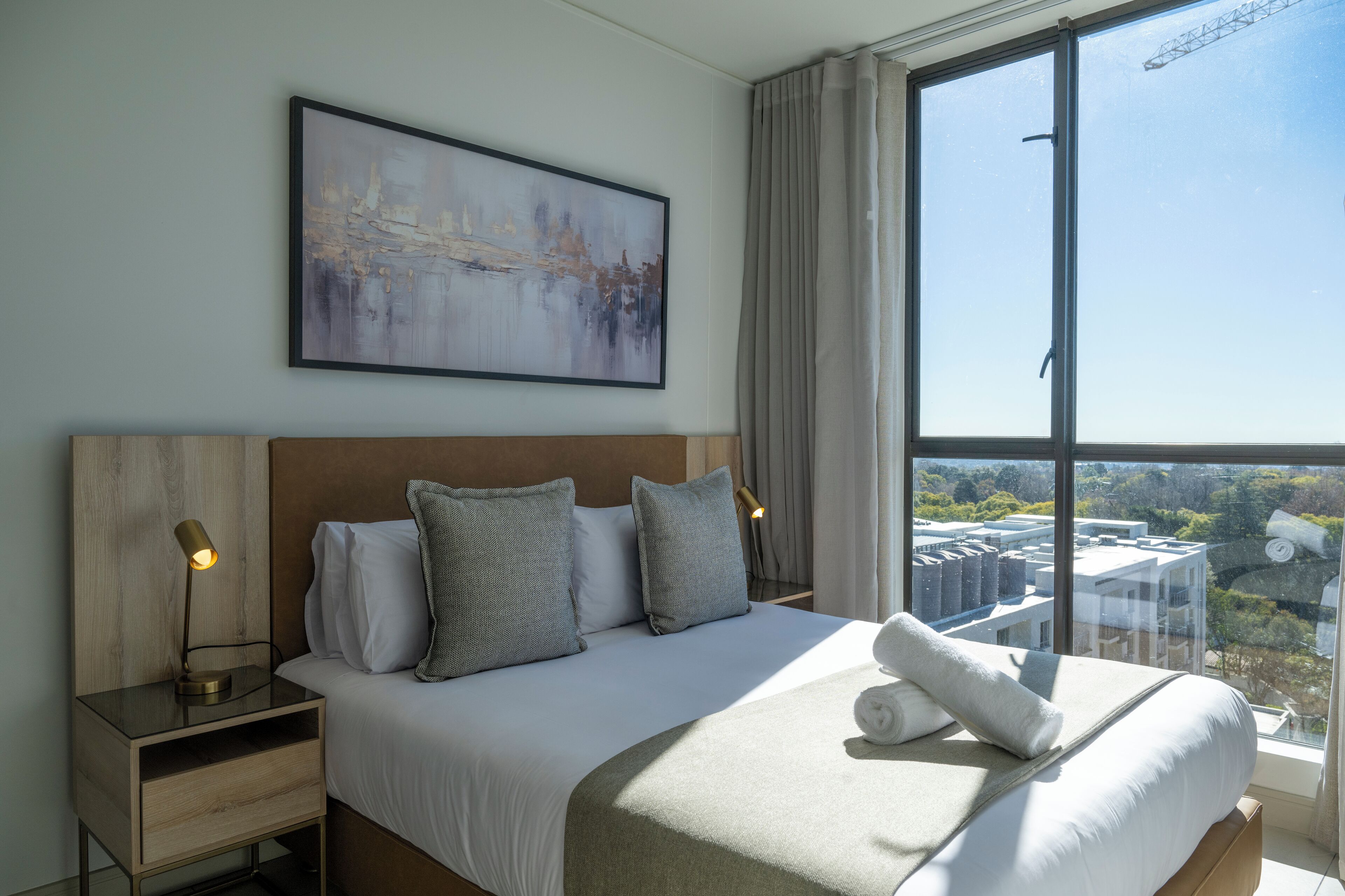 A modern hotel room with a large bed, a nightstand with a lamp, a painting on the wall, and a view of the city through the window.