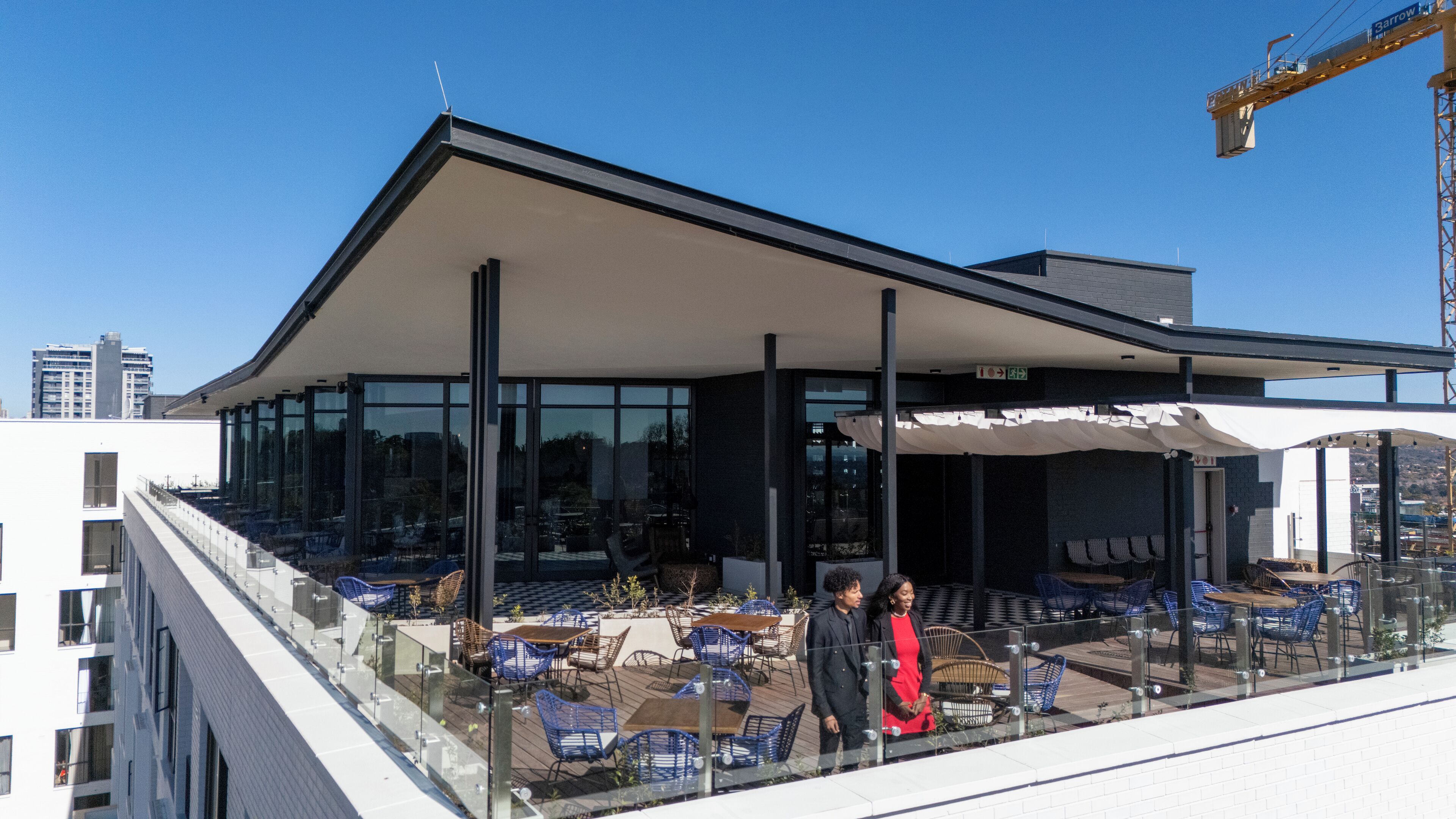 A modern building with a glass facade, outdoor seating area with blue chairs, and a covered patio.