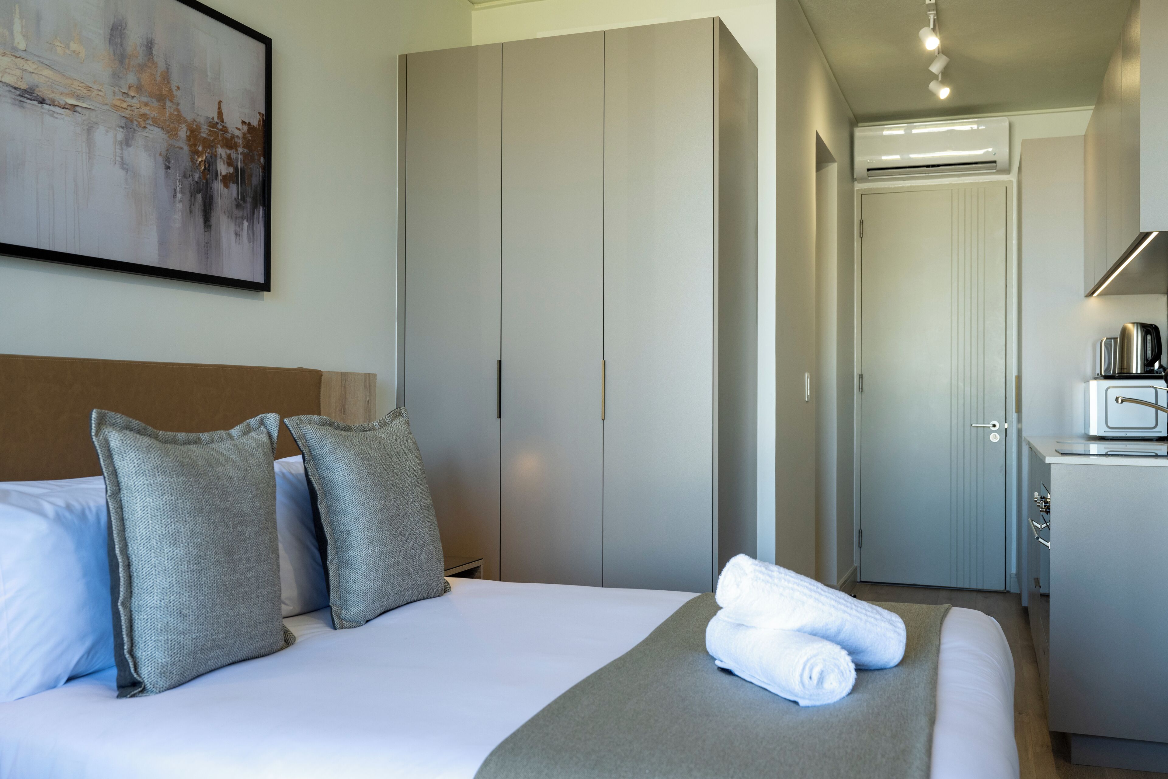 A hotel room with a bed, grey pillows, a grey throw blanket, and a neatly folded towel on the bed. There is a wardrobe with a mirror and a door leading to a bathroom.