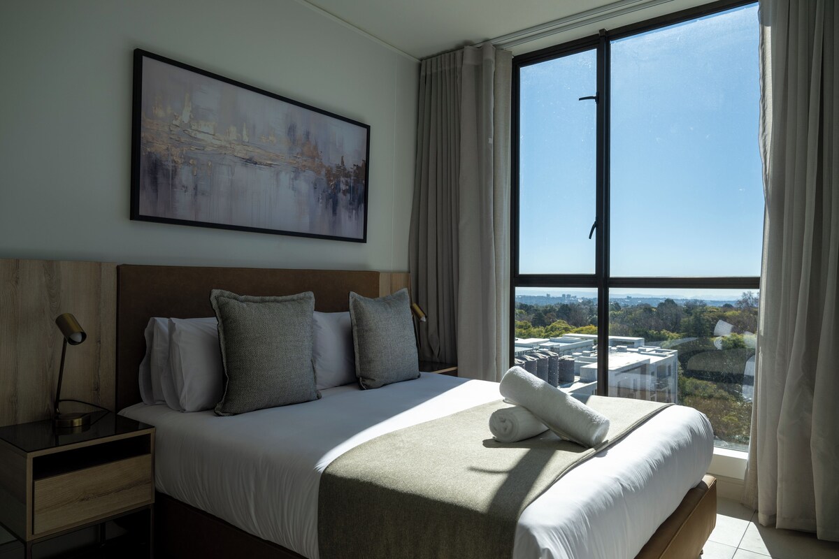 A hotel room with a large bed, a nightstand, a lamp, a painting on the wall, and a view of the city through a large window.