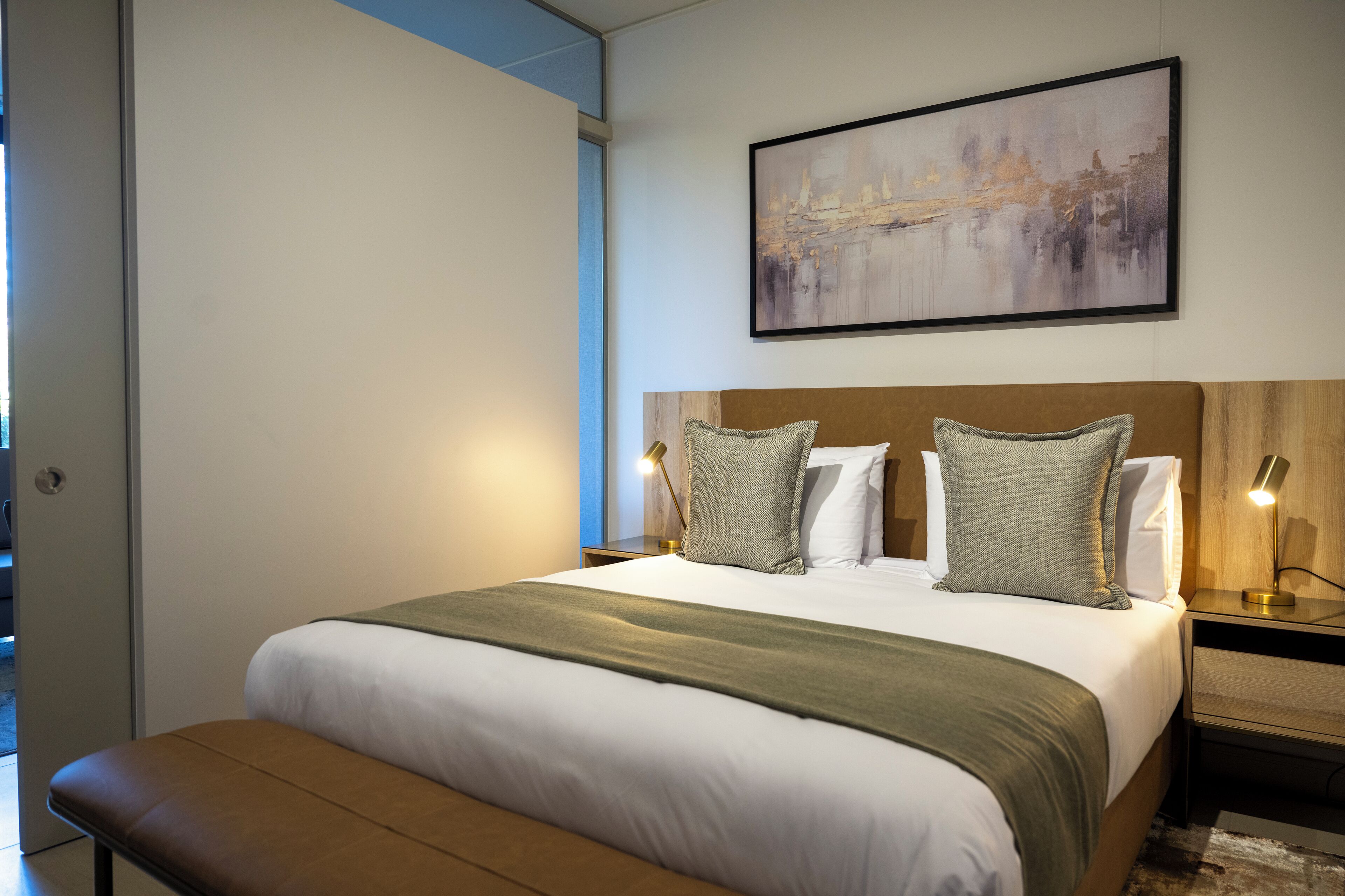 A neatly made bed with a green throw blanket, two bedside lamps, and a framed abstract painting above it.