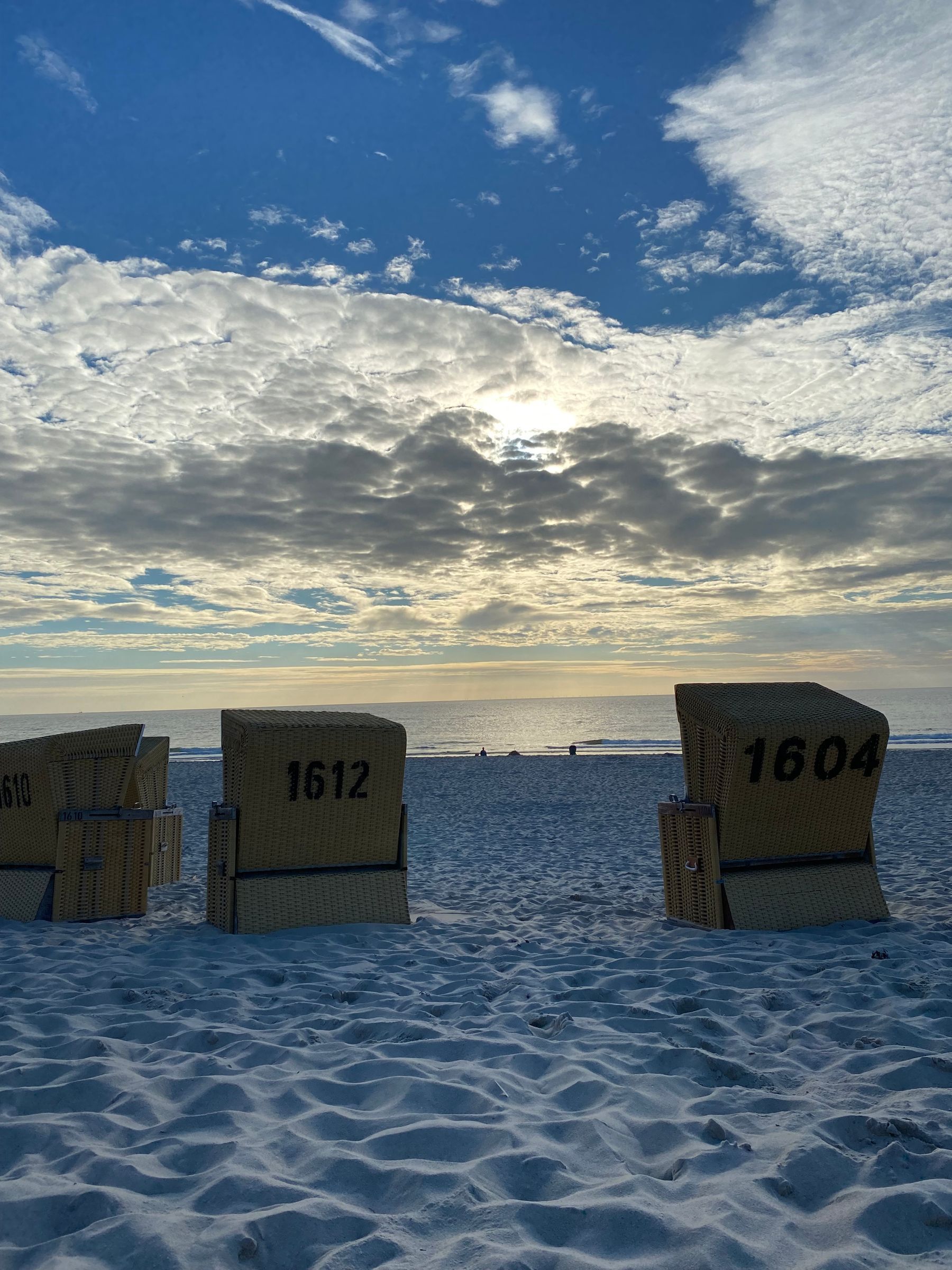Strandliegen mit den Nummern 1610, 1612 und 1604 an einem sandigen Strand unter einem bewölkten Himmel.