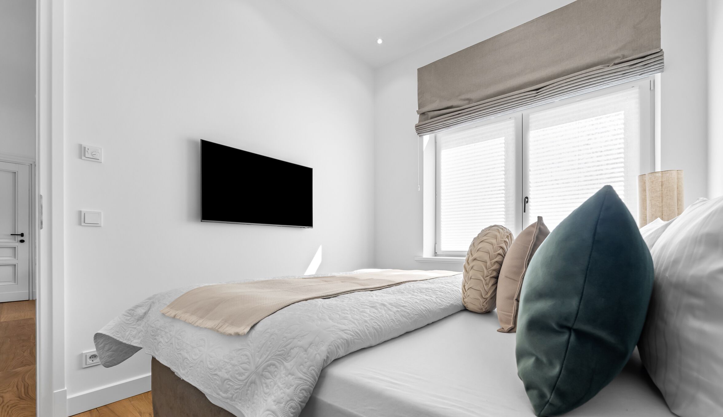 A modern bedroom with a bed, a flat-screen TV mounted on the wall, a window with blinds, and a bedside lamp.