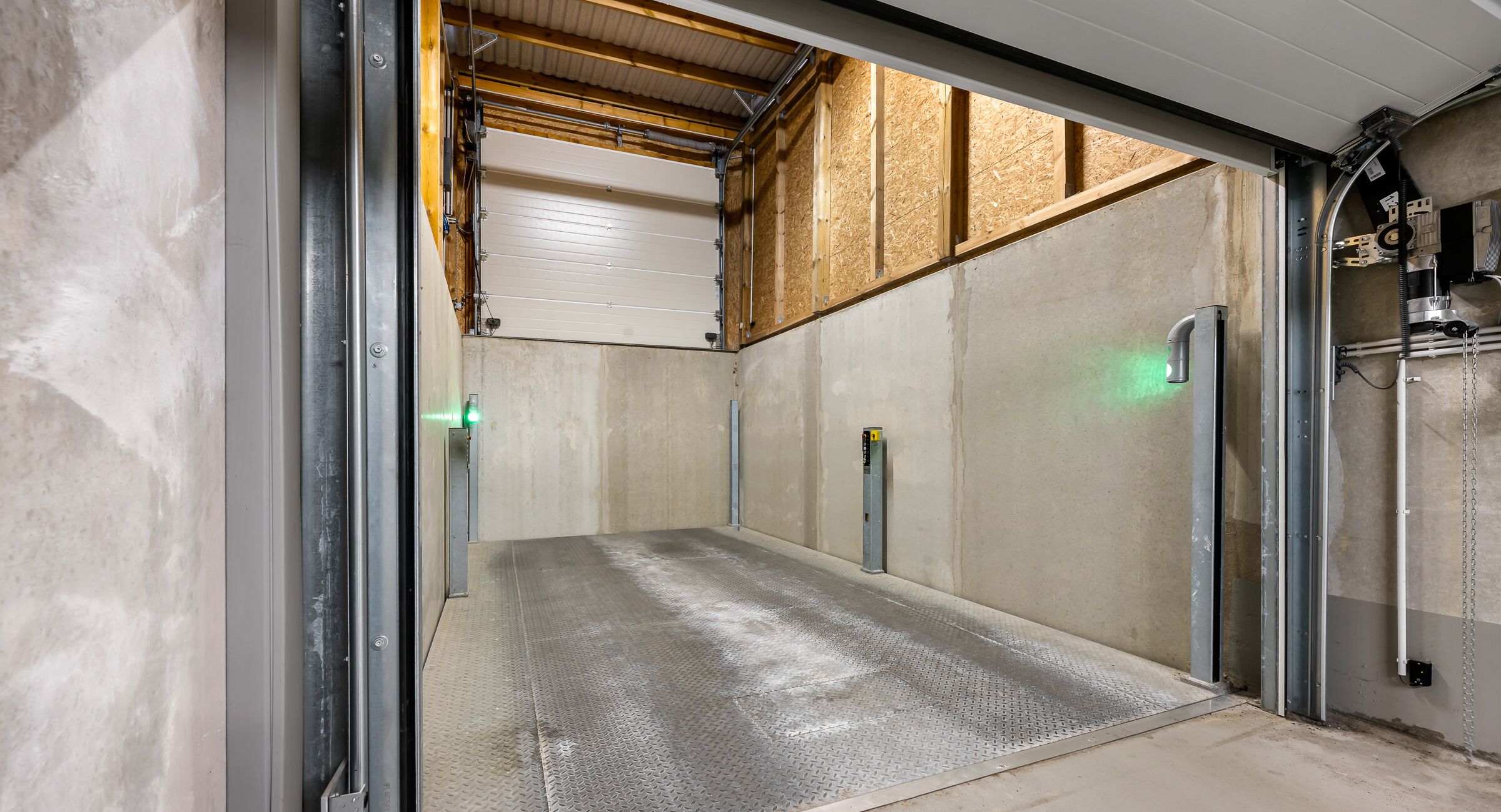 A garage with a metal ramp leading to a garage door, wooden framing on the ceiling, and a green exit sign.