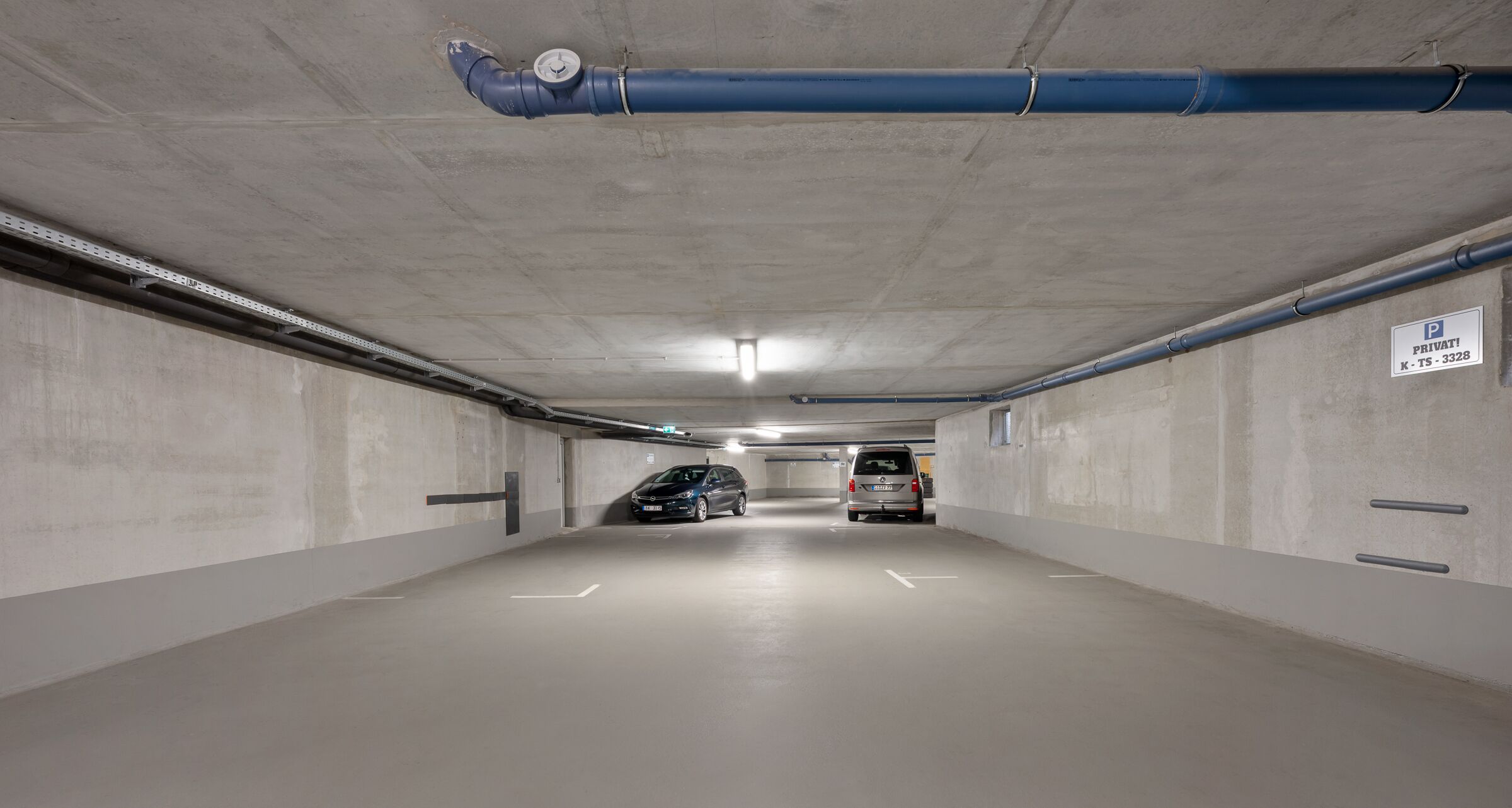 En parkeringskælder med betonvægge, et jævnt gulv og et loft med synlige rør. To biler er parkeret, og et skilt angiver privat parkering.