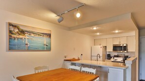 A compact kitchen with white cabinets, a wooden dining table, and a painting of a coastal scene.