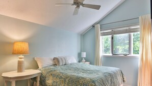 A bedroom with a bed, a bedside table, a lamp, a ceiling fan, a window with curtains, and a view of greenery outside.
