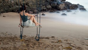 A person sitting on a swing on a sandy beach with rocks in the background. - Marleys Beach House (Talpe)