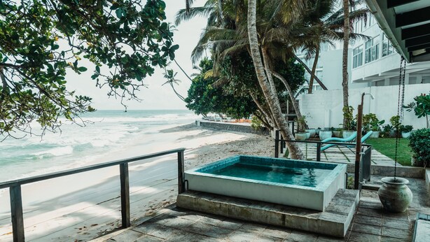A pool area with a view of the ocean, surrounded by palm trees and a white building. - Marleys Beach House (Talpe)