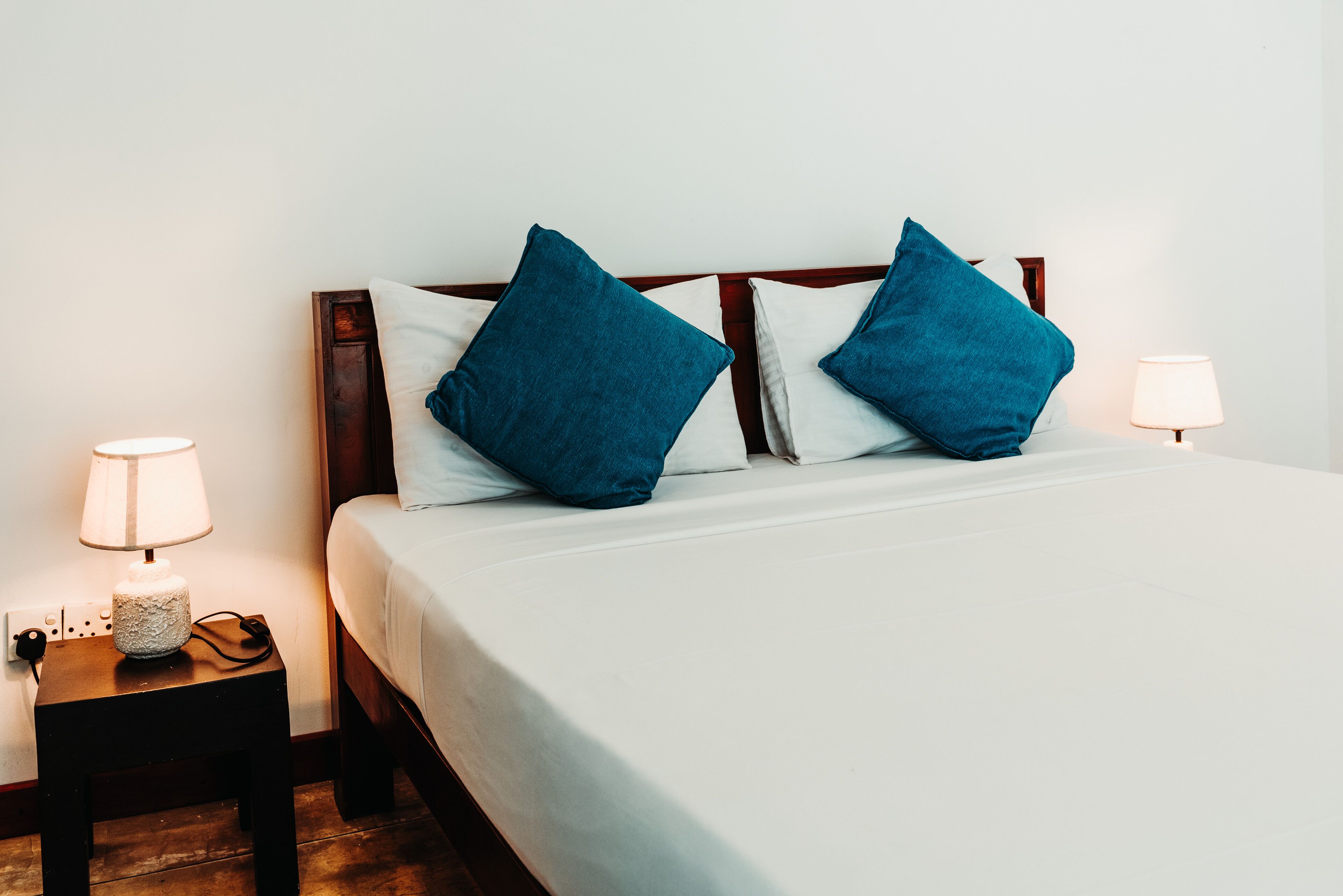A neatly made bed with white sheets and two blue pillows, flanked by two bedside tables with lamps.