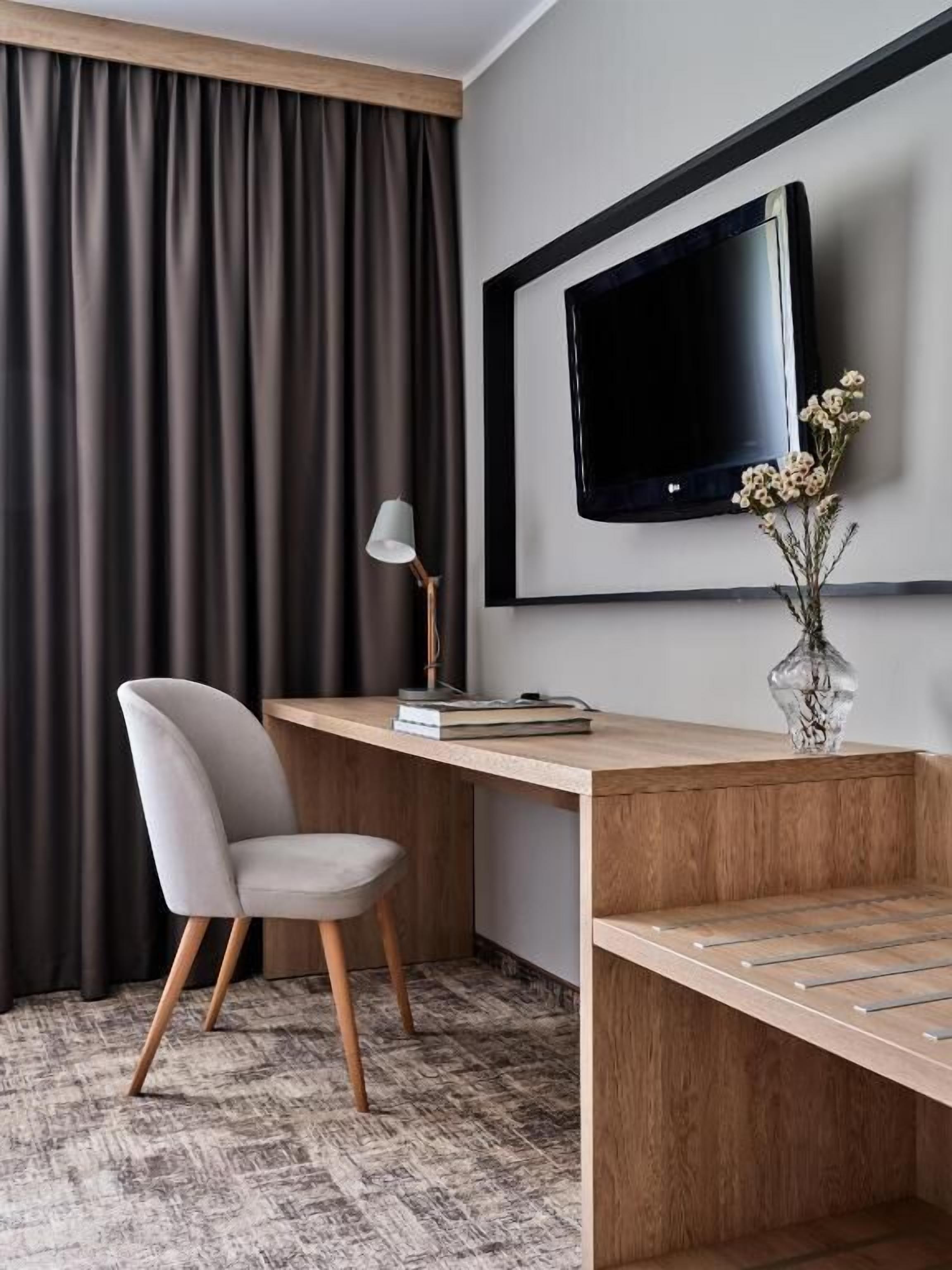 Ein modernes Zimmer mit Schreibtisch aus Holz, einem Sessel, einem an der Wand befestigten Fernseher und einer Vase mit Blumen auf dem Schreibtisch.