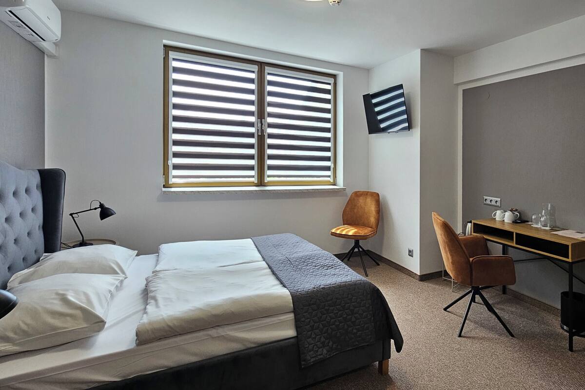 A bedroom with a bed, a desk, a chair, a window with blinds, and a mirror.