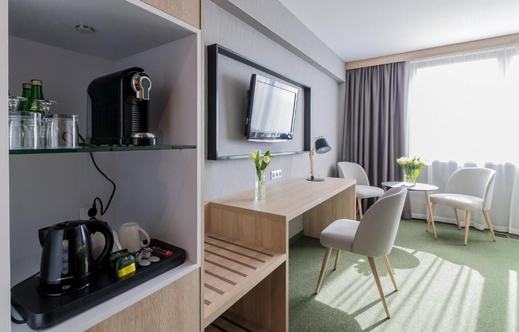 Habitación de hotel con escritorio, sillas, televisor, cafetera y estante con varios objetos.