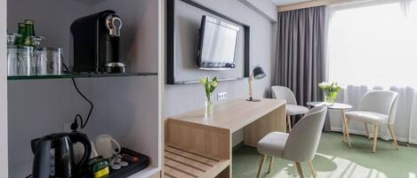 Habitación de hotel con escritorio, sillas, televisor, cafetera y estante con varios objetos.