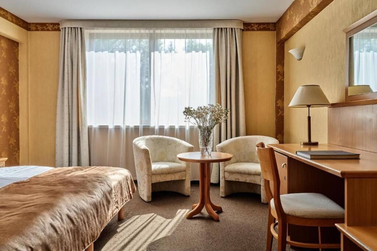 A hotel room with a bed, two armchairs, a small round table with a vase, a desk with a lamp, and a window with curtains.