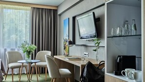 A modern hotel room with a wooden desk, two chairs, a television, a coffee machine, and a shelf with glasses and bottles. - Hotel Great Polonia Kraków Airport (Modlniczka)