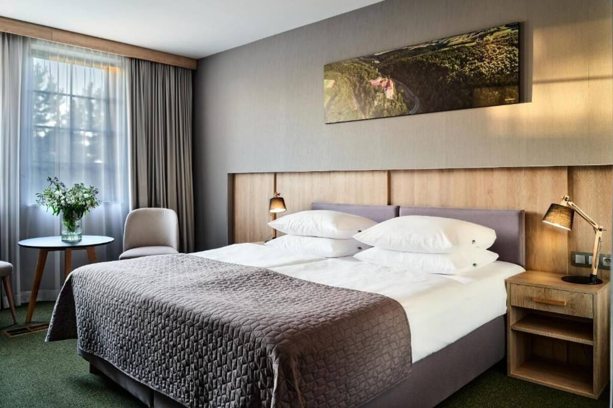 A hotel room with a large bed, a bedside table with a lamp, a chair, a small table with a vase, and a painting on the wall.