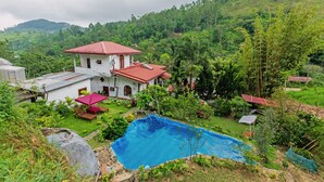 A white building with a red roof, a swimming pool, and lush greenery surrounding it.
