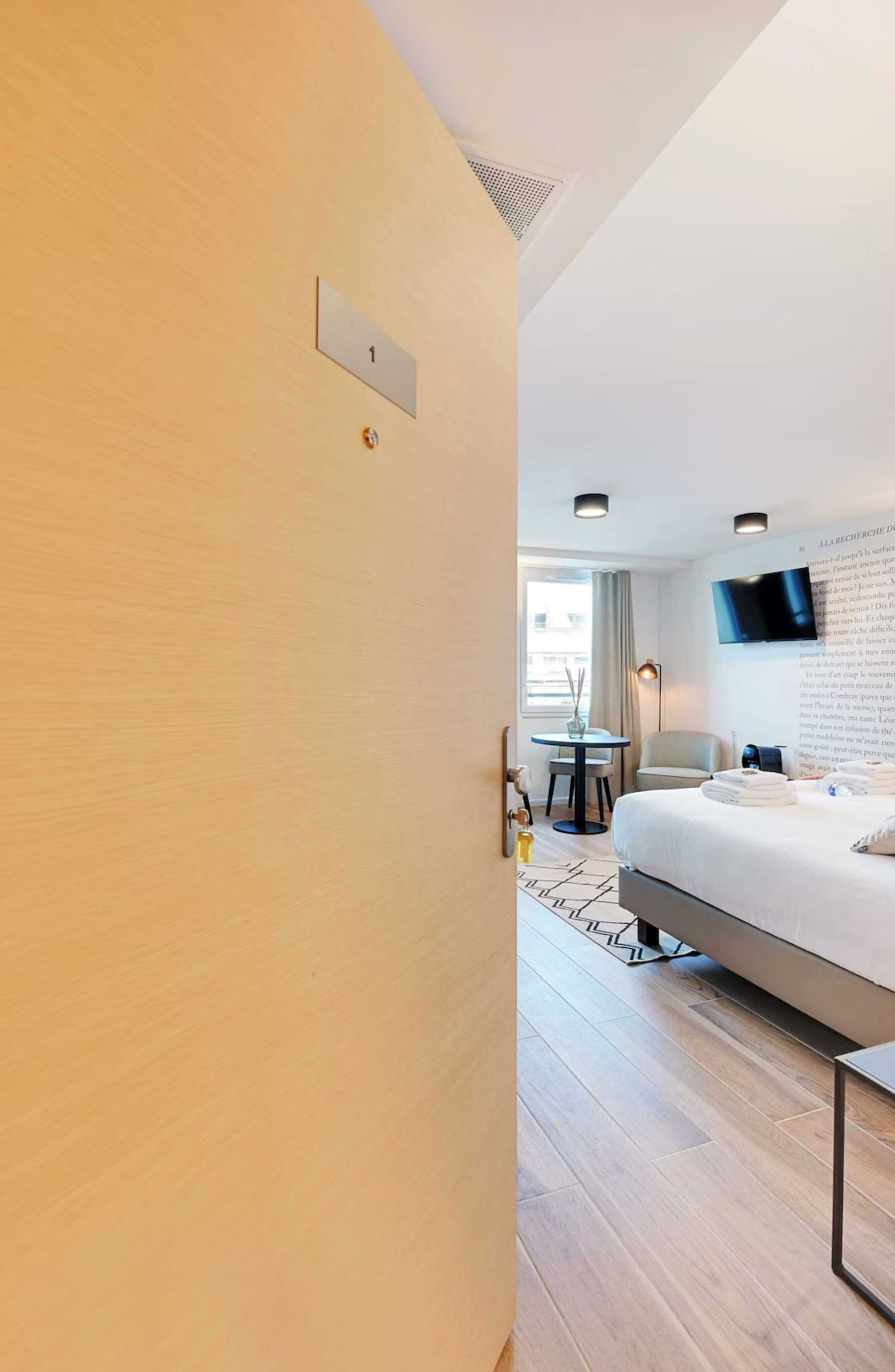 A modern hotel room with a bed, a desk, a chair, a TV mounted on the wall, and a window with curtains.