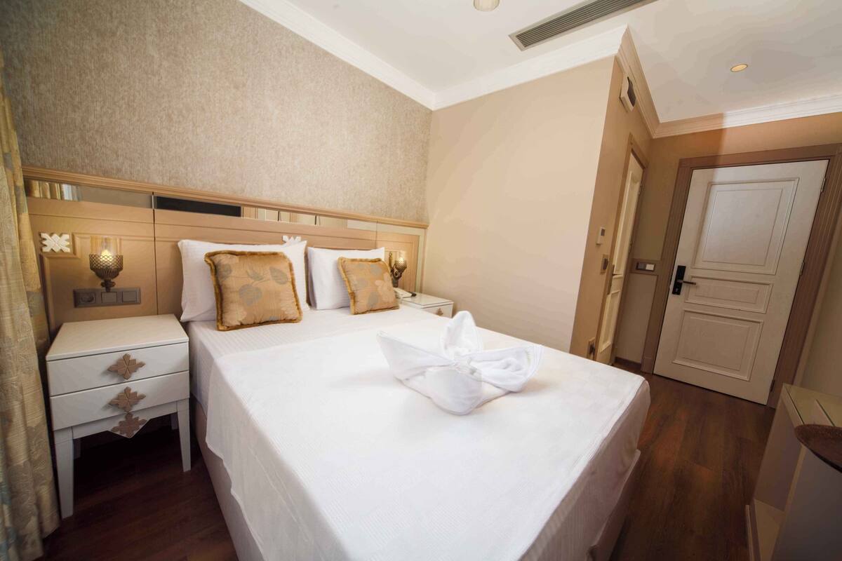 A hotel room with a neatly made bed, a bedside table with a lamp, and a door to the right.
