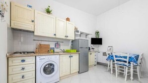 Cocina compacta con armarios blancos, lavadora, refrigerador y comedor con mesa y sillas.