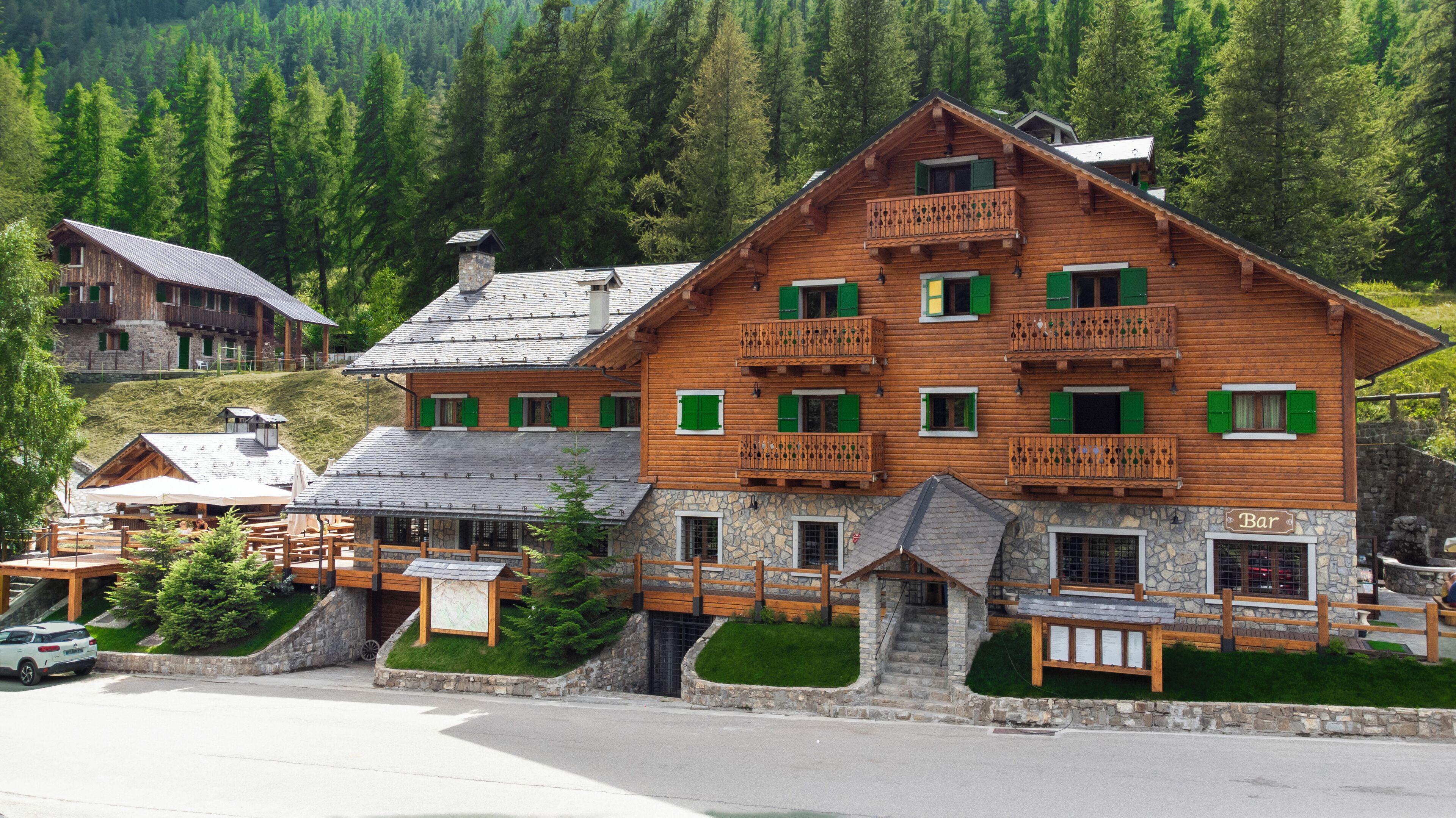 A wooden chalet with multiple balconies and a stone facade, surrounded by lush greenery and a mountainous backdrop.