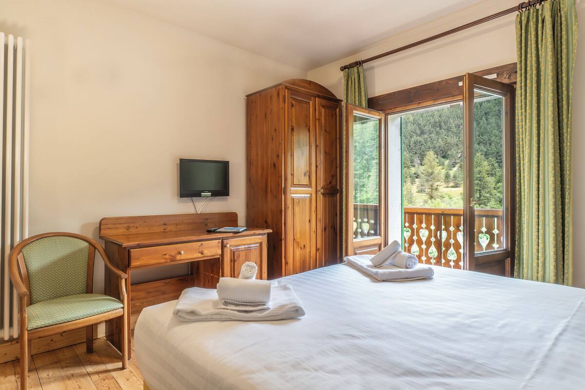 A hotel room with a bed, a desk, a chair, a TV, and a balcony with a view of trees.