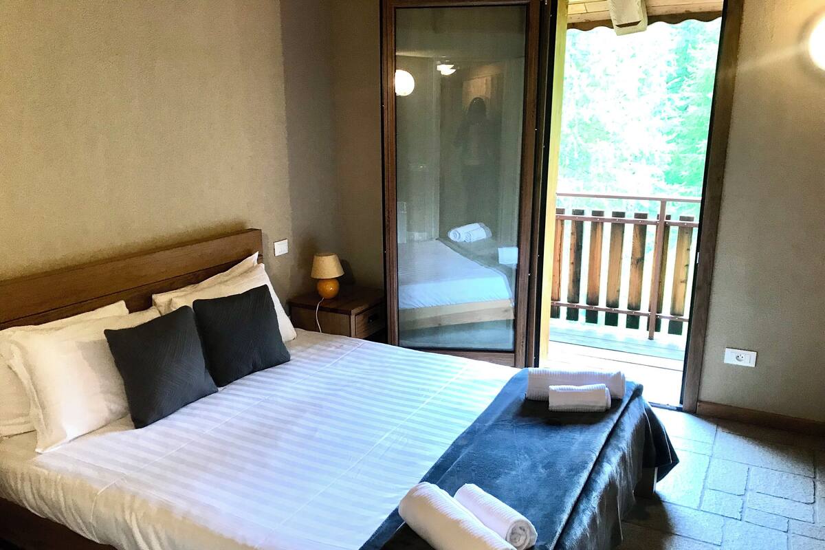 A neatly made bed with white and dark pillows, a blue bedspread, and two rolled towels on the side.