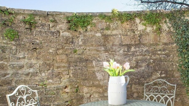 Ein Metall-Garnitur mit Tisch und Stühlen vor einer Steinmauer, mit einer weißen Vase mit Blumen.