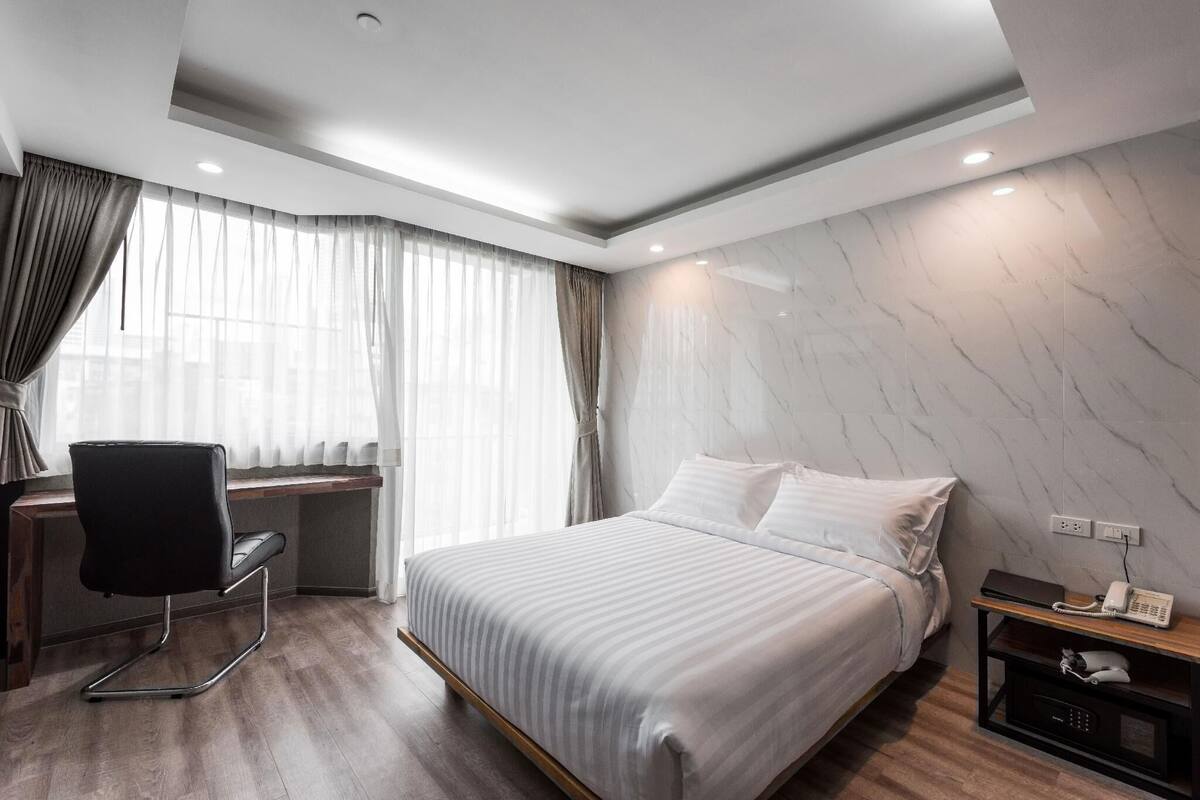 Ein modernes Hotelzimmer mit Bett, Schreibtisch mit Stuhl, Nachttisch mit Telefon und einem Fenster mit Vorhängen.