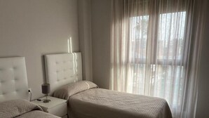 Ett hotellrum med två sängar, ett nattduksbord, en lampa och ett stort fönster med tunna, genomskinliga gardiner.
