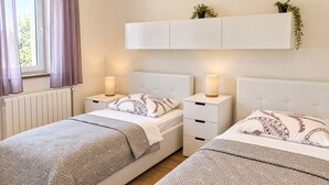 A hotel room with two beds, bedside tables, a radiator, and a window with curtains.