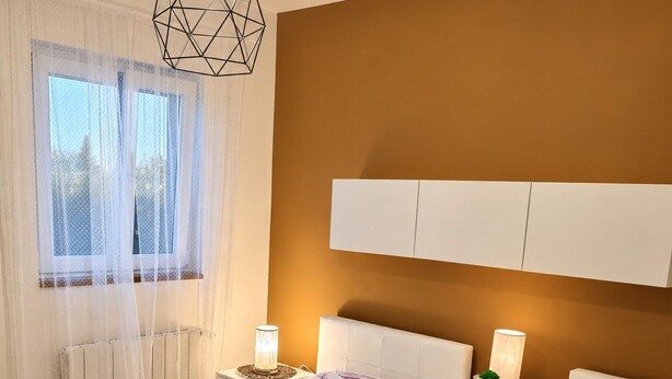 A bedroom with two beds, a window with sheer curtains, bedside lamps, and a geometric light fixture.