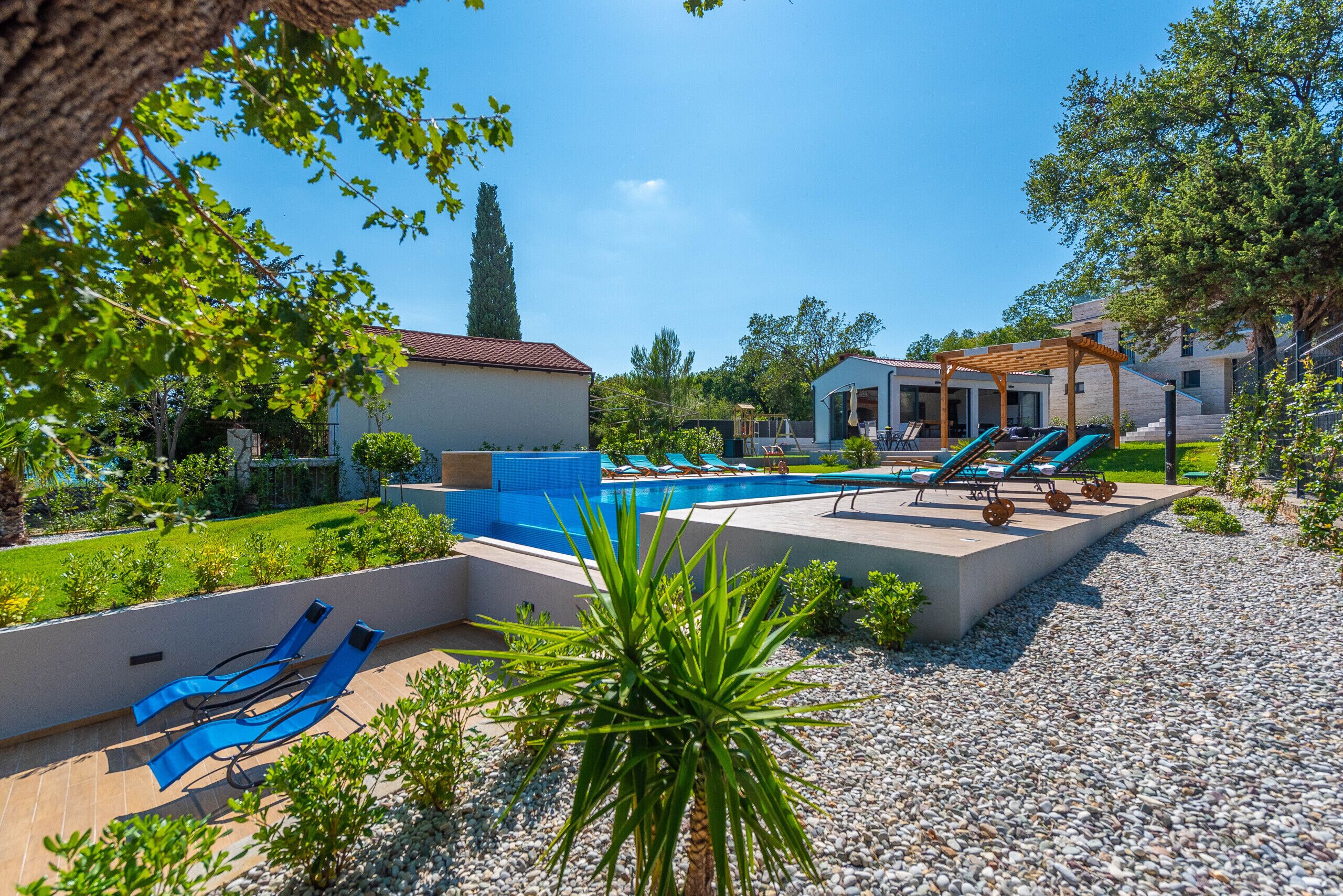 A pool area with lounge chairs and a pergola, surrounded by greenery and a pebble path.