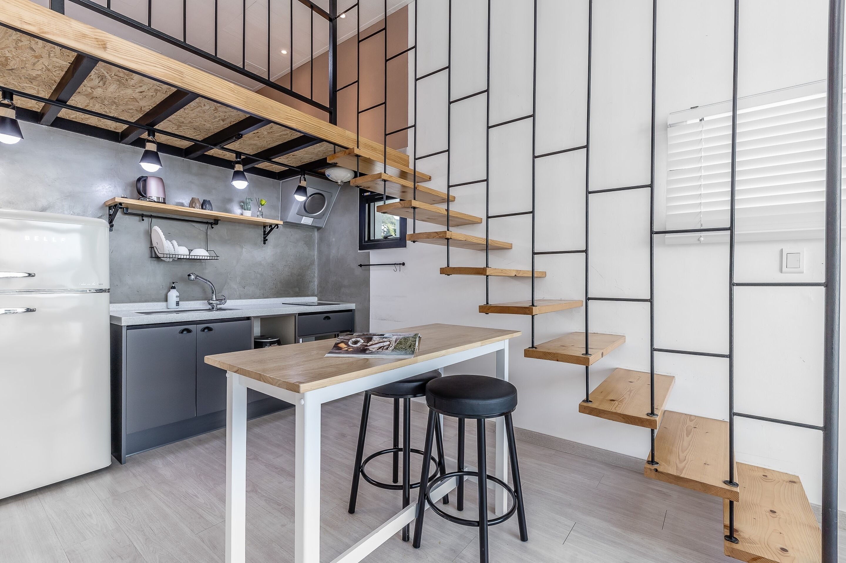 A modern kitchen with a wooden table, bar stools, and a staircase with wooden steps.