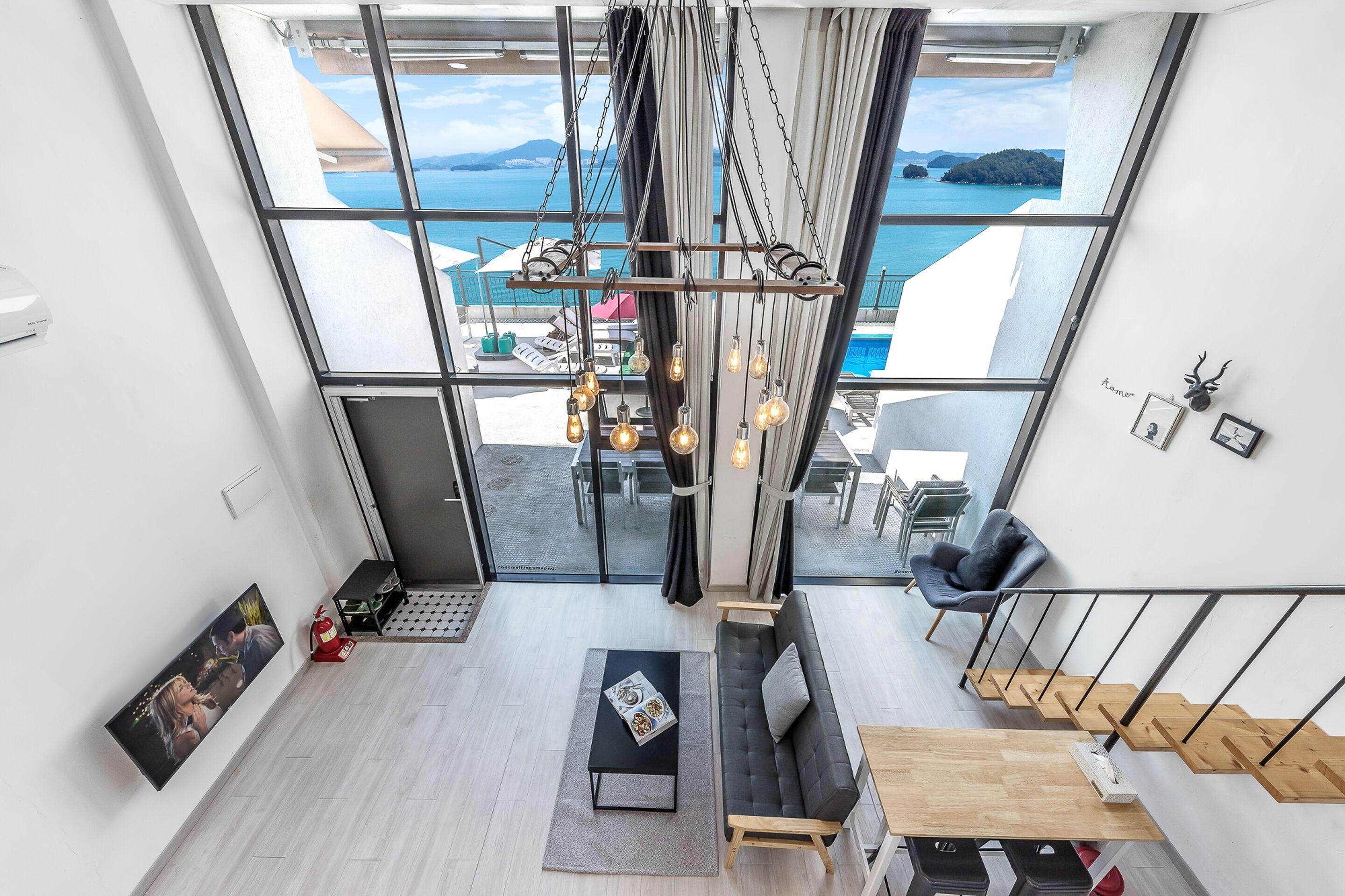 A modern interior with a large window overlooking a scenic ocean view, a black sofa, a wooden table, and a hanging light fixture.