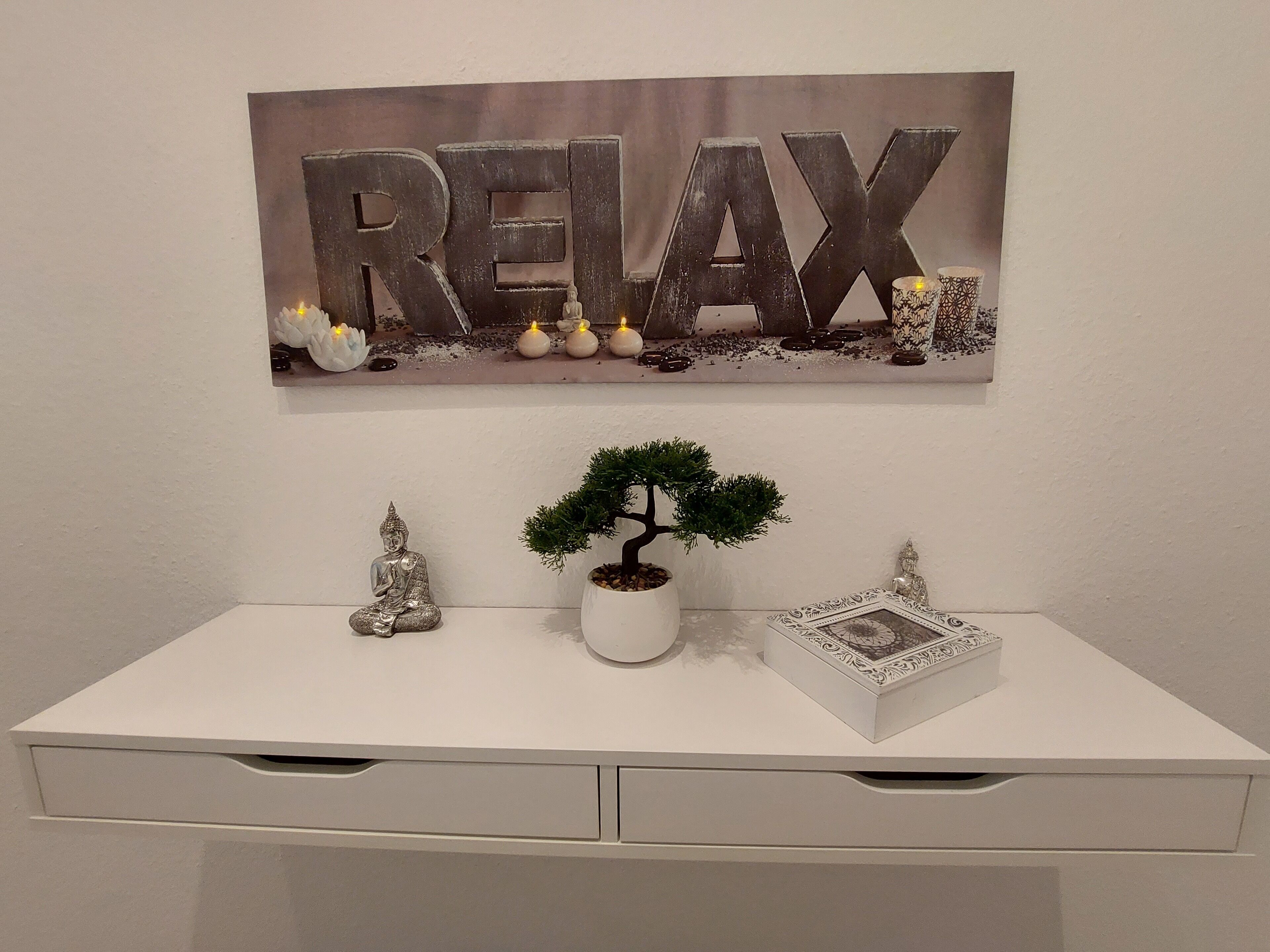 A white desk with a bonsai tree, two Buddha statues, and a framed word "RELAX" above it.
