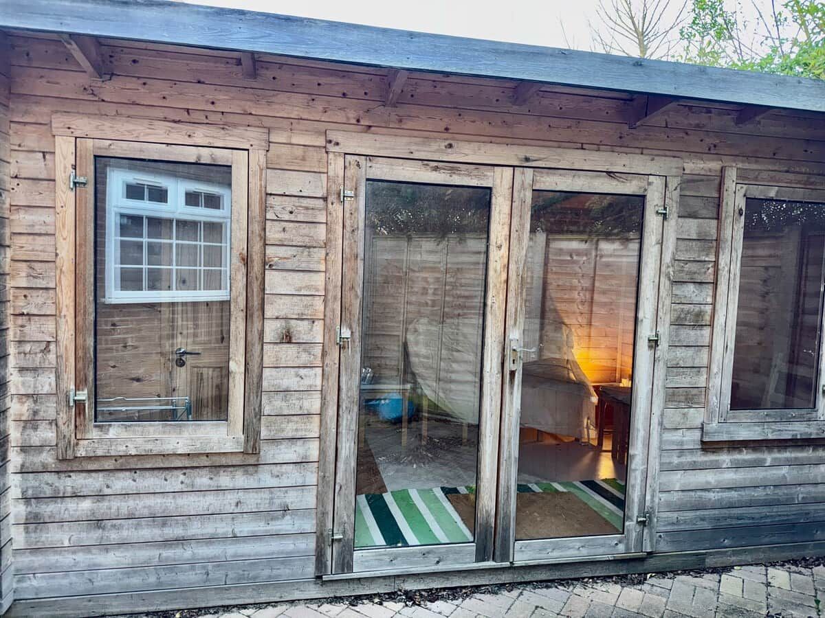 A wooden cabin with a sliding glass door, a window, and a visible interior with a bed and a lamp.