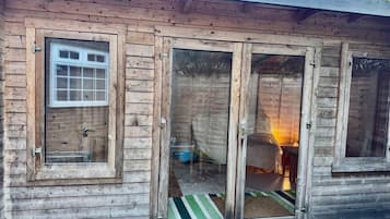 A wooden cabin with a sliding glass door, a window, and a visible interior with a bed and a lamp.