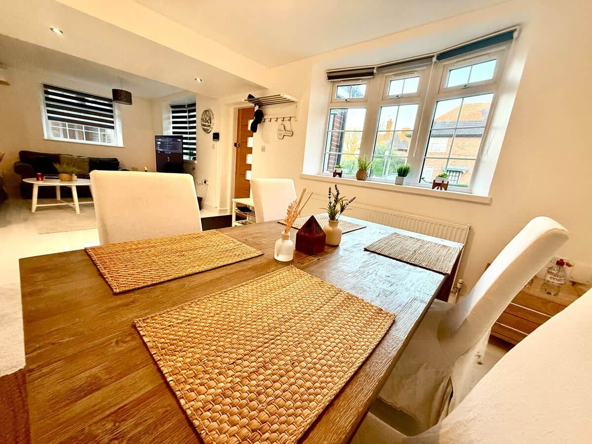 A dining area with a wooden table, woven placemats, white chairs, and a window with blinds.
